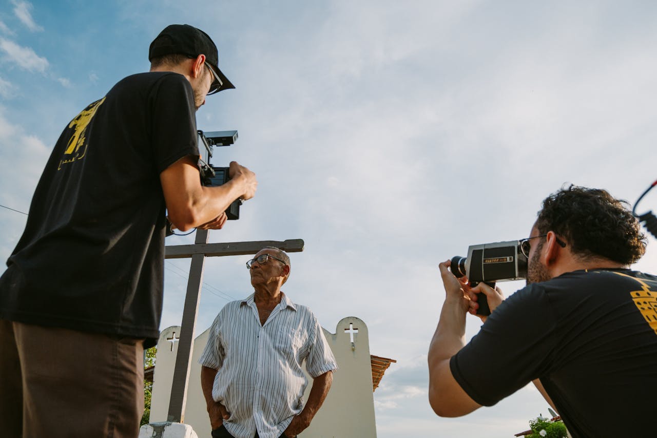 Two filmmakers document an elderly man in an outdoor setting, capturing the moment with professional cameras.